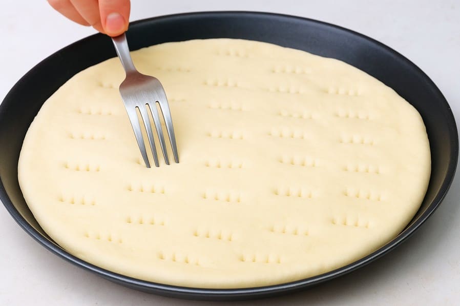 बेला हुआ डो प्लेट पर रखा है और कटे वाले चमच से हल्के छेद किए जा रहे हैं