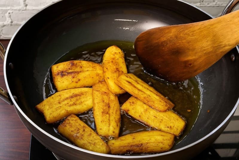 लोहे की कड़ाही में कच्चे केले के टुकड़े फ्राई करते हुए – दोनों तरफ सुनहरा होने तक भूनते हुए दृश्य